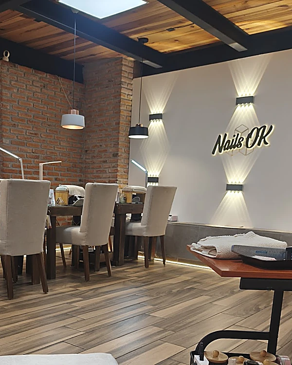 Interior de Nails OK: el letrero iluminado sobre el muro blanco, la columna de ladrillo, lámparas colgantes y las mesas de manicura con sillas blancas