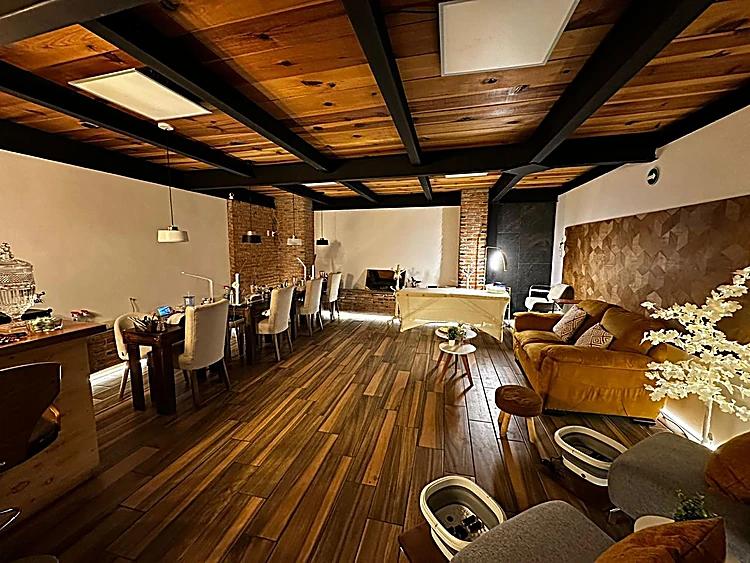 Vista del salón con techo de madera y vigas negras, piso de duela y, al fondo a la derecha, la sala de pies con un sillón mostaza
